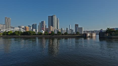 a panoramic view of tokyo's modern skyline, featuring numerous high-rise buildings and a bridge, seen across a calm body of water.