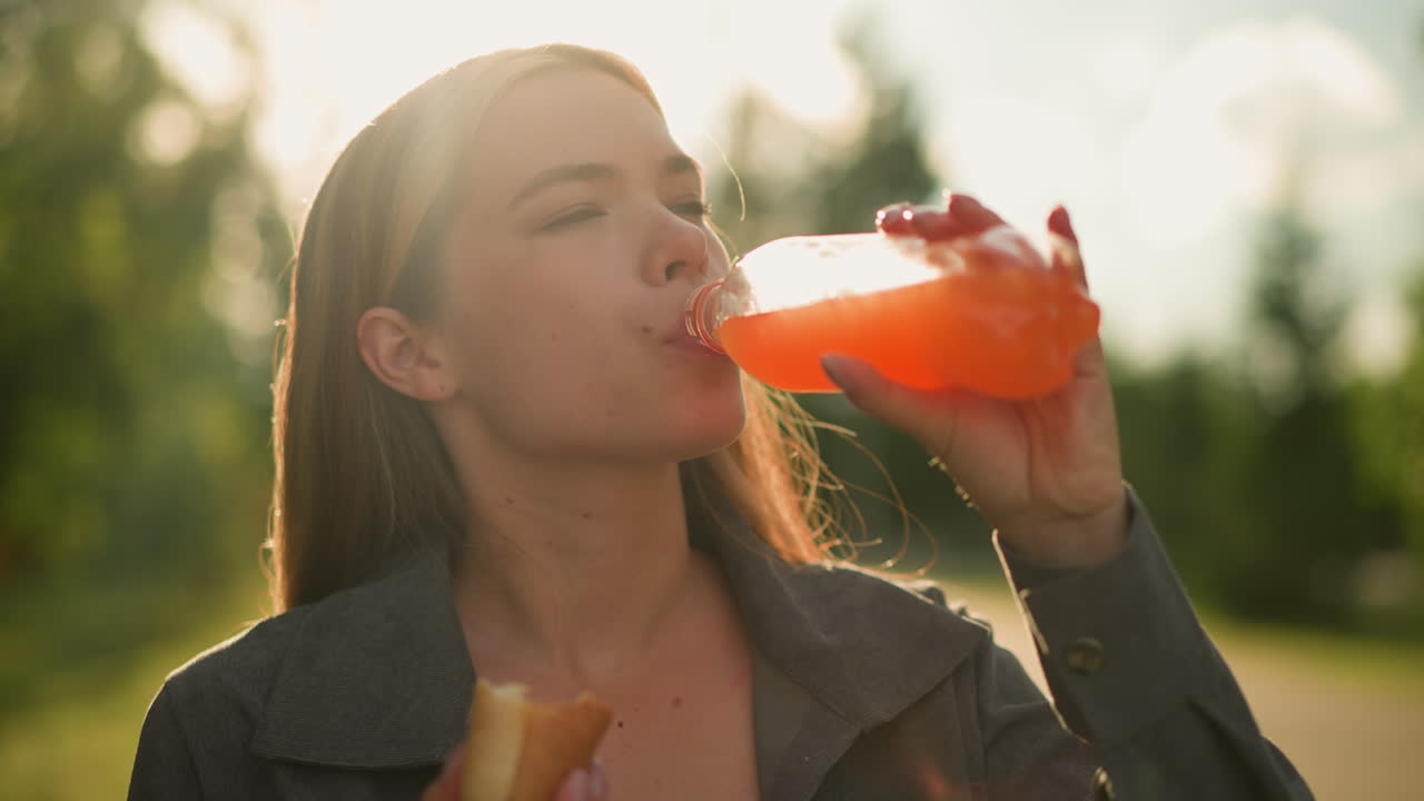 Lady in grey clothing sips from juice bottle while chewing a sausage roll in her hand, the outdoor scene features a blurred background with glowing sunlight filtering through trees
