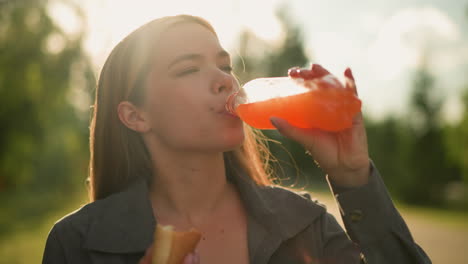 una dama con ropa gris toma sorbos de una botella de jugo mientras mastica un rollo de salchichas en su mano, la escena al aire libre presenta un fondo borroso con la luz del sol brillante filtrándose a través de los árboles