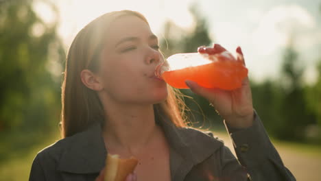 lady in grey clothing sips from juice bottle while chewing a sausage roll in her hand, the outdoor scene features a blurred background with glowing sunlight filtering through trees