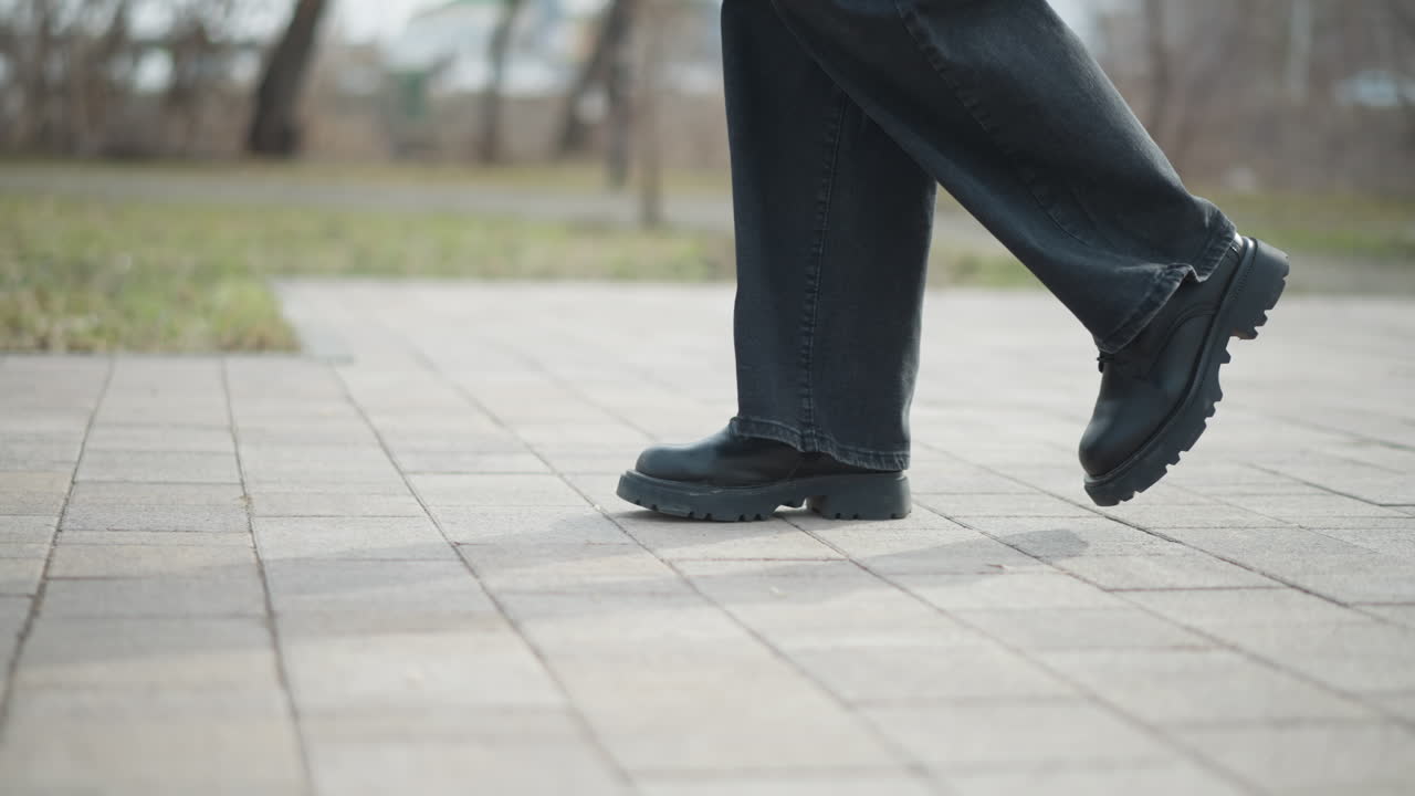 Close-up of legs in motion showing black shoes and dark pants walking on paved outdoor surface during cold season, capturing moment of stride with emphasis on movement, texture, and balance in urban park