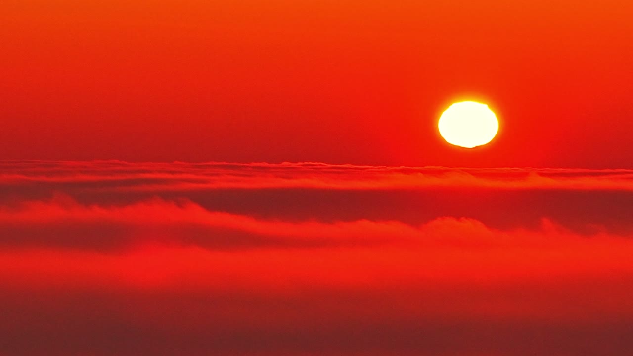 Drone view of the sun above a sea of clouds at dusk. Deep red tones fill the sky as sunlight fades along the horizon, creating a dramatic, abstract background with strong atmospheric mood