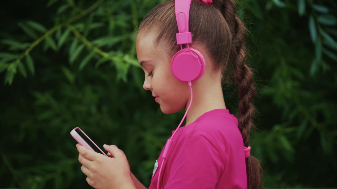 A Young Girl Enjoys Her Favorite Music, Engrossed in Her Smartphone While Wearing Stylish Pink Headphones in a Lush Green Setting