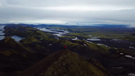 aerial drone landscape view of one person wearing a red jacket, standing on top of a mountain, in the iceland highlands