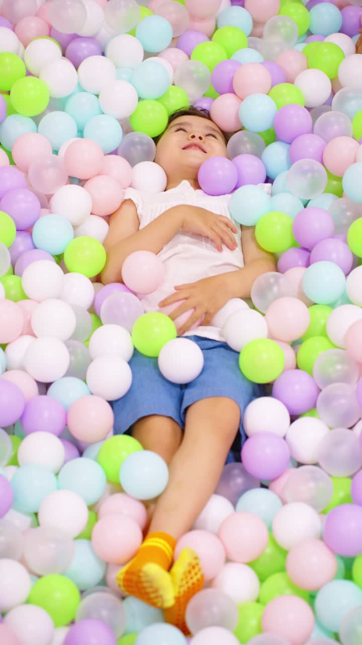 A joyful young child, lies peacefully amidst a vibrant sea of pastel-colored balls in an indoor play area, surrounded by soft shades of pink, purple, green, white, and blue