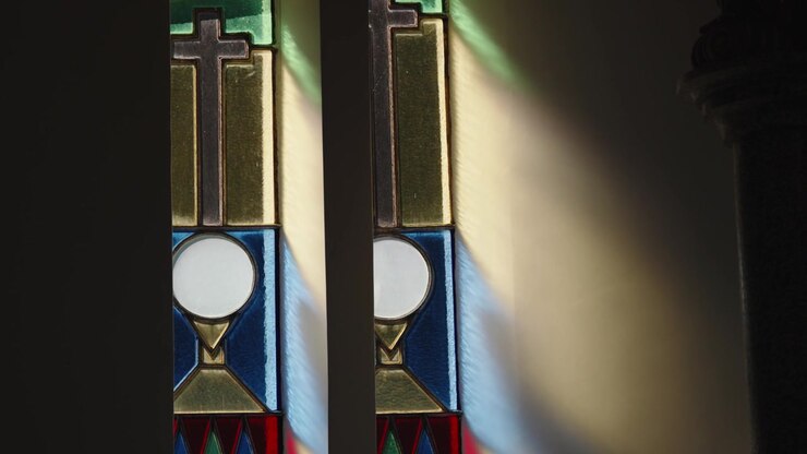 Two narrow stained glass windows showing a cross, a white communion host and a gold chalice in blue and yellow panes, light falling on a dark church wall.