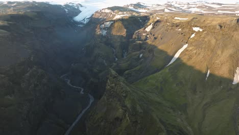 drone shot of a dramatic icelandic canyon with a winding river, towering cliffs, and a glacier in the background under sunlight