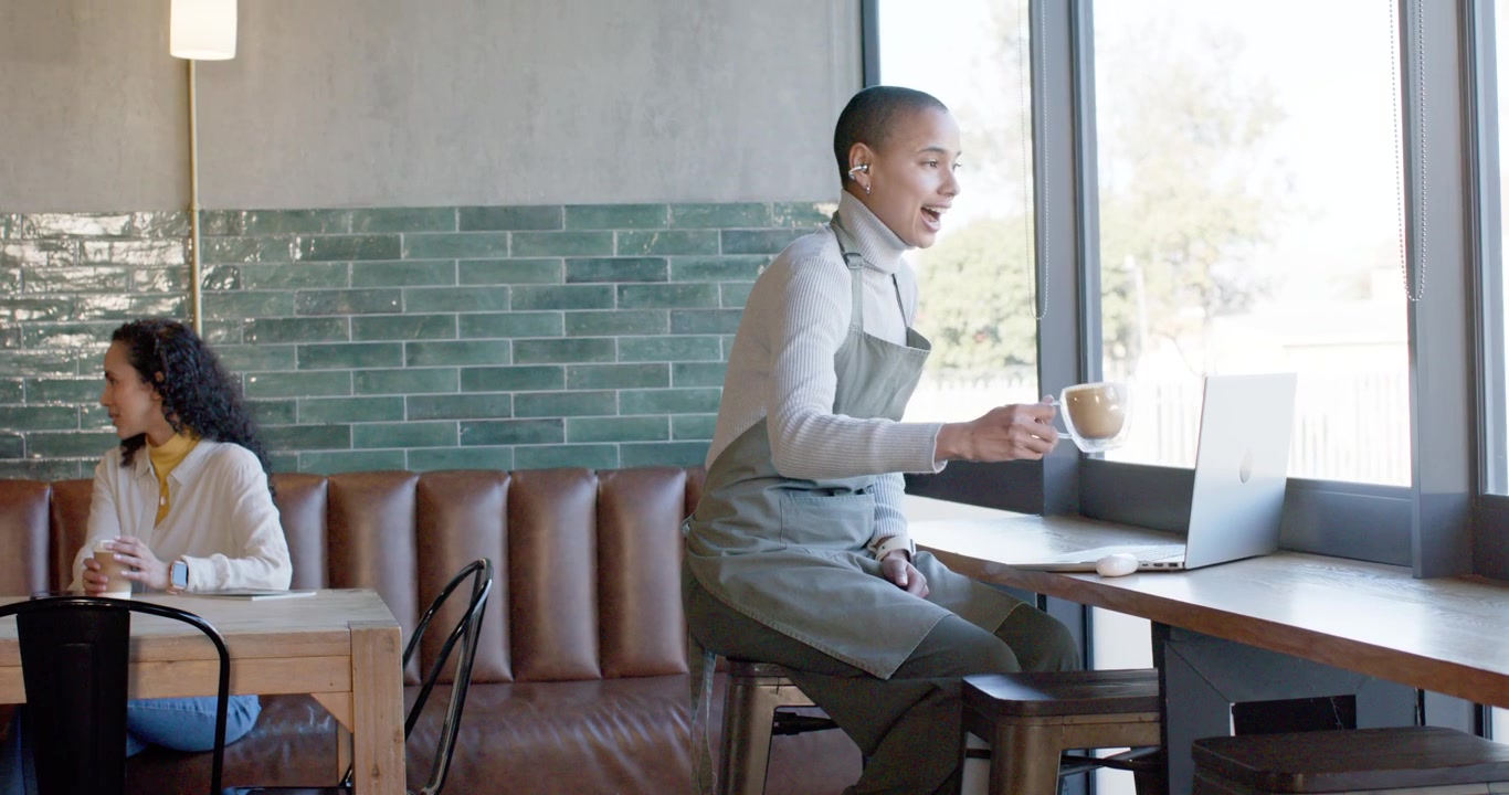 African American barista wearing green apron, typing on laptop at window counter