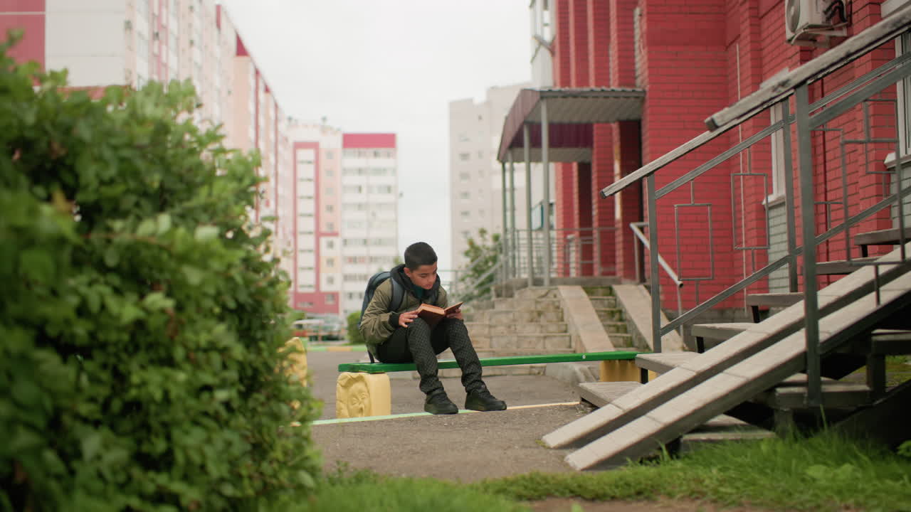 바람에 흔들리는 식물 옆 빨간 벽돌 건물 근처 초록색 벤치에 앉아 책을 읽으며 페이지를 넘기는 아이 (어깨에는 백팩을 메고 있어요)