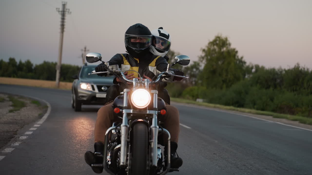 Motorcycle rider with passenger wearing helmets makes sharp bend on paved road while cars follow on other lanes, glowing headlight illuminating asphalt during evening travel