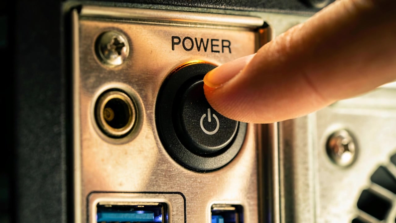 Approaching adult fingertip pressing power button on computer at desk, powering system, LED glowing