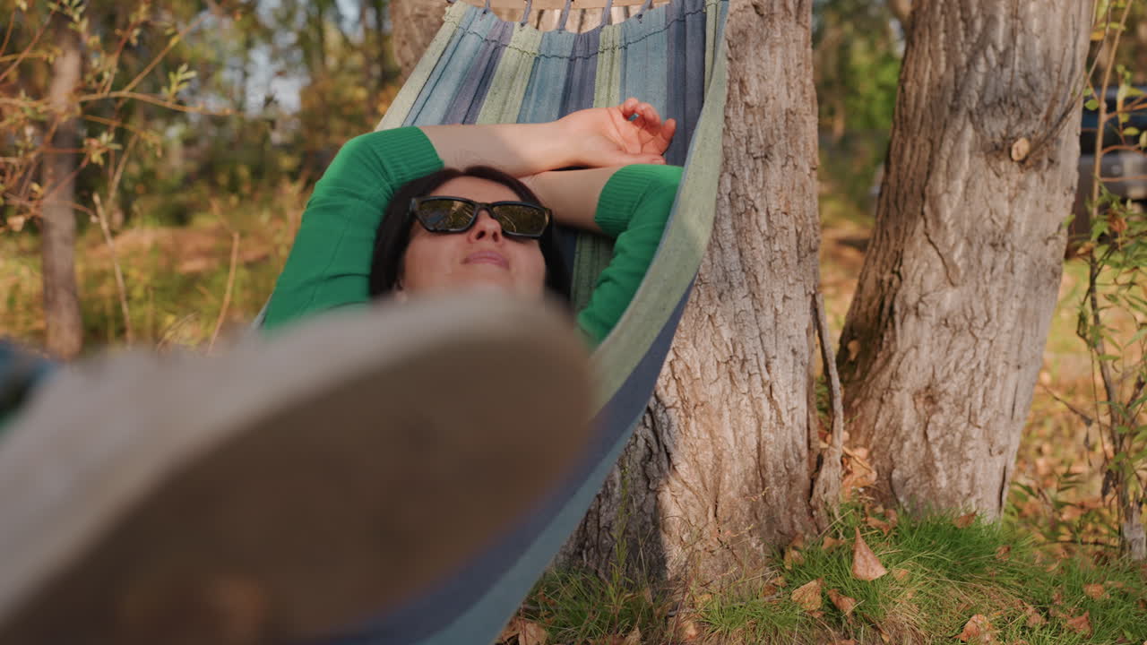 Primeiro plano aproximado de tênis de camping em rede visto de baixo, com a sola do tênis preenchendo o quadro. Pessoa reclinada em suéter verde, fundo de floresta embaçado, corda e tronco visíveis, movimento relaxado, luz solar suave e grama de outono.