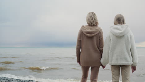 Back-view:-Mother-and-daughter-look-at-the-ocean,-where-storm-clouds-are-in-the-distance.-Standing-on-the-shore,-holding-hands