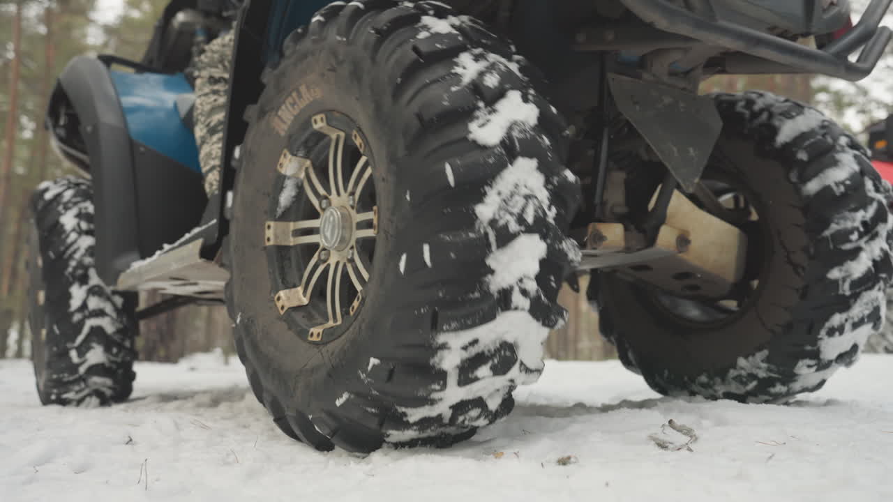 In una foresta innevata, primo piano della pedana di un quad e dei pneumatici coperti di neve, con i pantaloni mimetici del pilota appoggiati sul piolo. Inquadrature posteriori dal basso mostrano il battistrada robusto e le sospensioni, pini sullo sfondo, con una luce fredda e nitida.