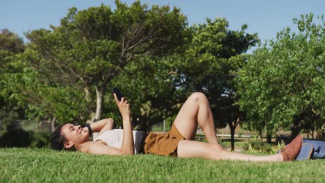 Mujer-Afroamericana-Tumbada-En-El-Césped-Usando-Un-Teléfono-Inteligente-En-El-Parque