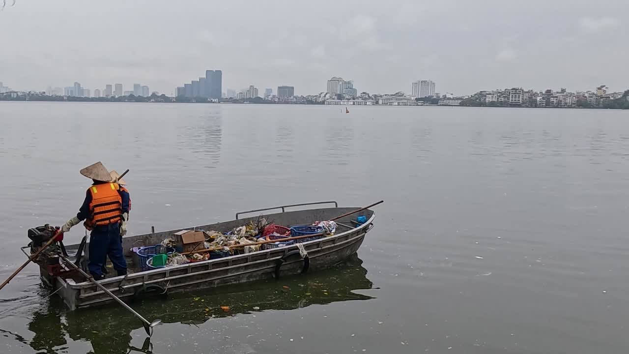 Two volunteers in life vests gather debris from a river using a small boat, with a cityscape in the background.