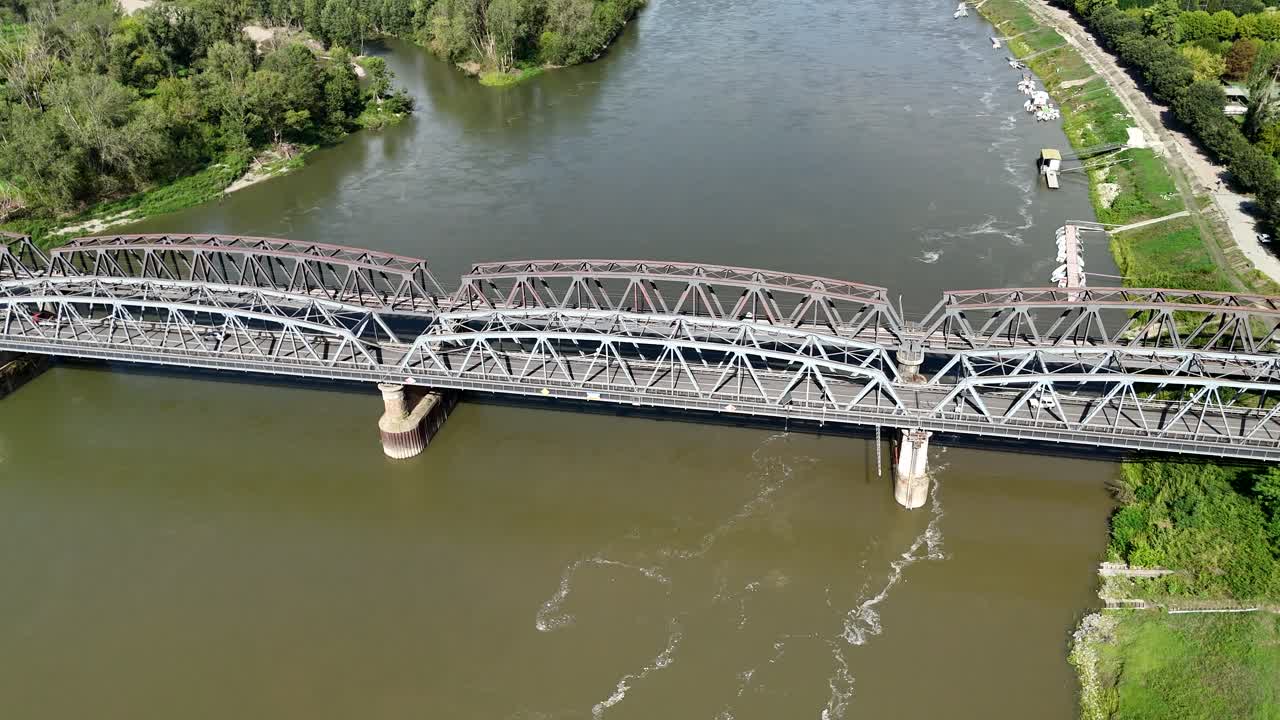 drone pulls away and camera tilting down over iron bridge between cremona - lombardy and castelvetro piacentino . emilia romagna over Po river, with riverbanks lined by greenery