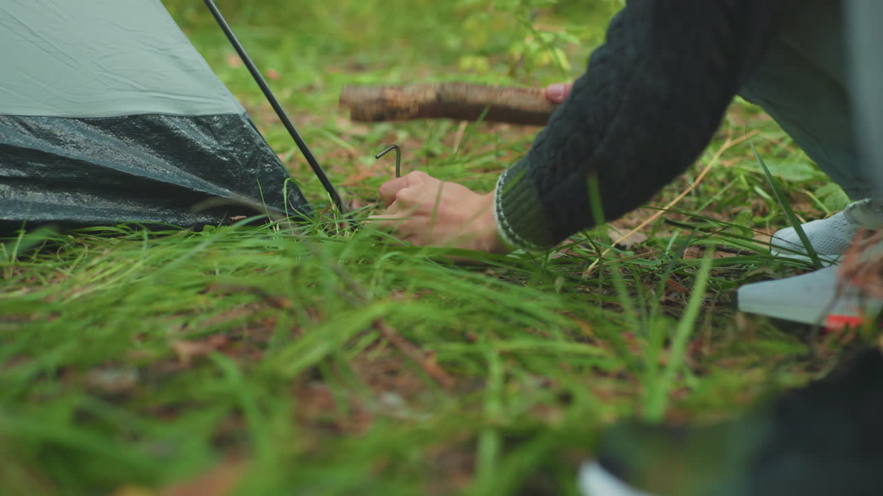 close up low leg level view of companion standing beside crouched camper as he hammers tent peg into soft forest ground amid grass pine needles securing camp canvas shelter