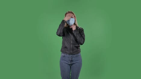 portrait of beautiful young woman puts on medical mask. blonde with loose hair in a black jacket and jeans on a green screen in the studio. health protection corona virus concept