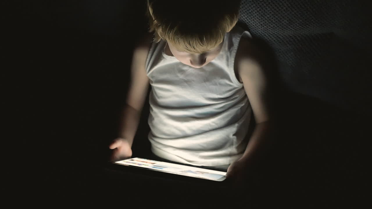 밤에 침대 시트로 덮인 온라인 비디오를 보는 태블릿을 사용하는 어린 소년