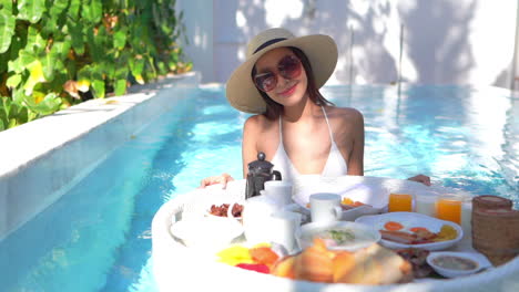 pretty exotic woman with breakfast plate for two in swimming pool on sunny morning at tropical holiday, full frame