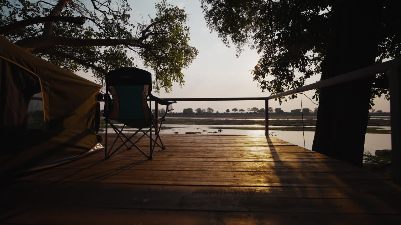 Slow dolly movement revealing a Gonarezhou campsite at sunset, with tent, deck, river view, and backlight. Serene atmosphere, captured in slow motion