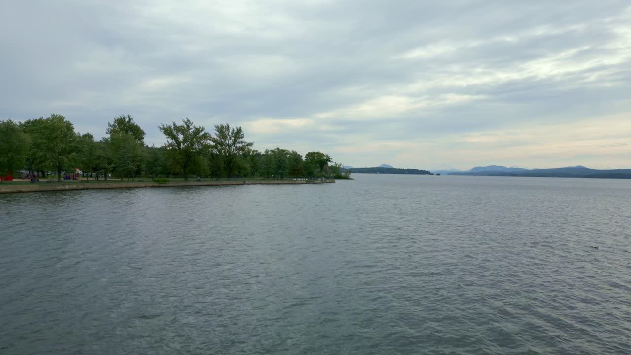 Lake Memphremagog in Magog, Canada, drone pushes towards over calm waters. Lush shoreline and distant mountains under soft cloudy sky during dusk.