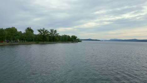 lake memphremagog in magog, canada, drone pushes towards over calm waters. lush shoreline and distant mountains under soft cloudy sky during dusk.