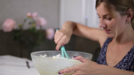 Young-woman-prepares-dough-mixing-ingredients-in-the-the-bowl-using-whisk-in-the-kitchen-and-looking-in-the-camera.-Homemade