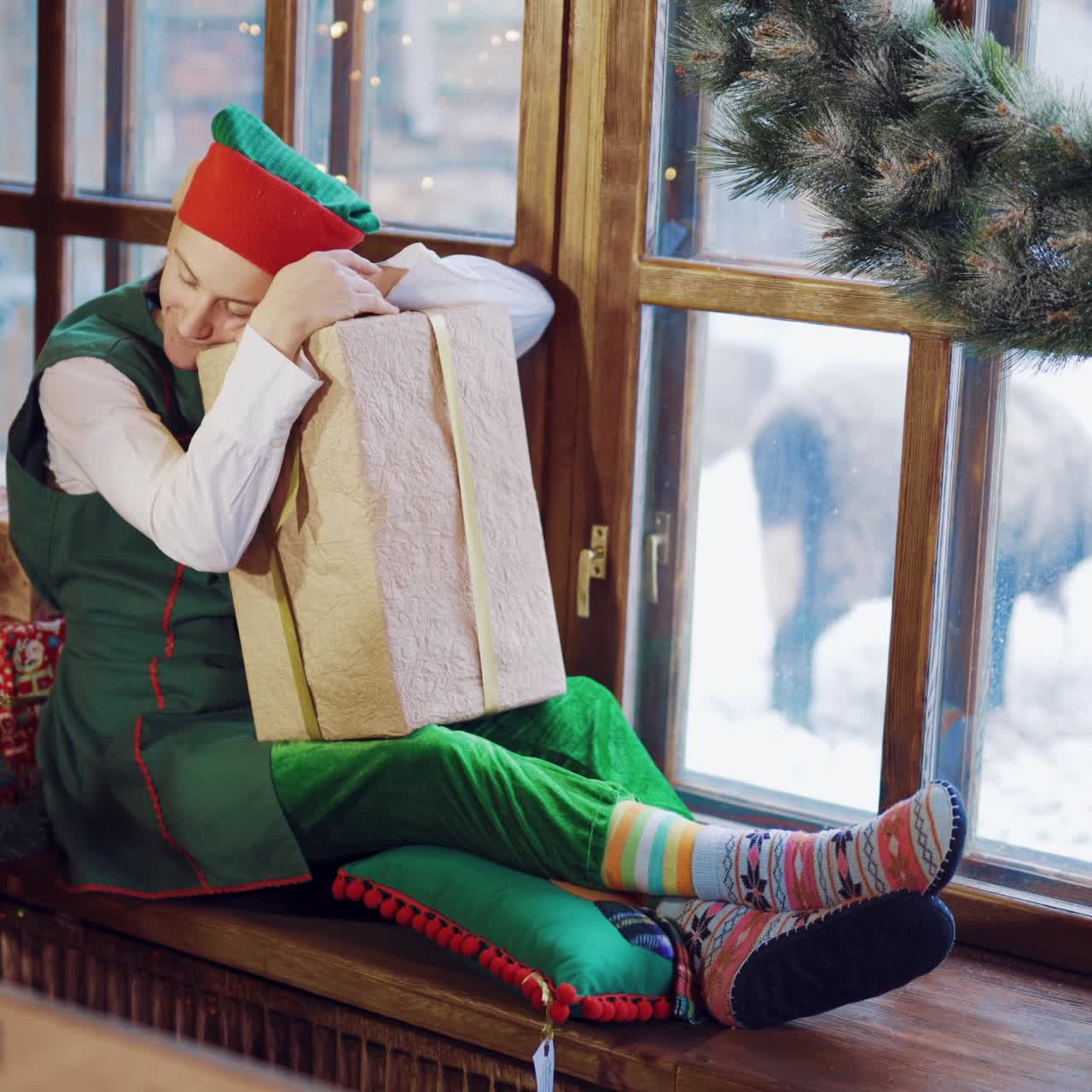 Tired elf with long ears is sitting on the windowsill with a gift in his hand on the background of the window near the Christmas tree in anticipation of the New Year. Magic creatures
