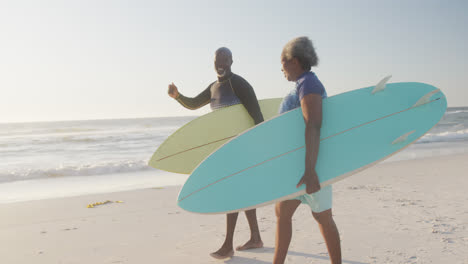 happy senior african american couple walking and holding surfboards at beach, in slow motion