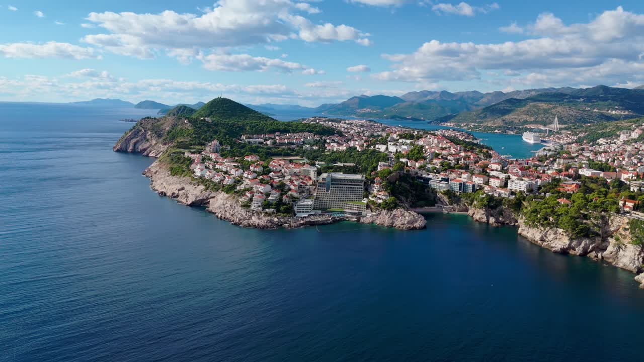 Wide-angle aerial drone flies toward Dubrovnik’s coastal town, revealing terracotta rooftops, winding streets, and the deep blue Adriatic Sea, with the coastline and surrounding landscape