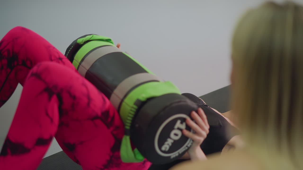 Donna su tappetino che esegue un ponte per i glutei con sandbag, attivazione mirata dei fianchi in uno studio calmo; coach che osserva la forma, leggings rosa, respirazione controllata, ripetizioni lente, enfasi sulla stabilità pelvica e sul core.
