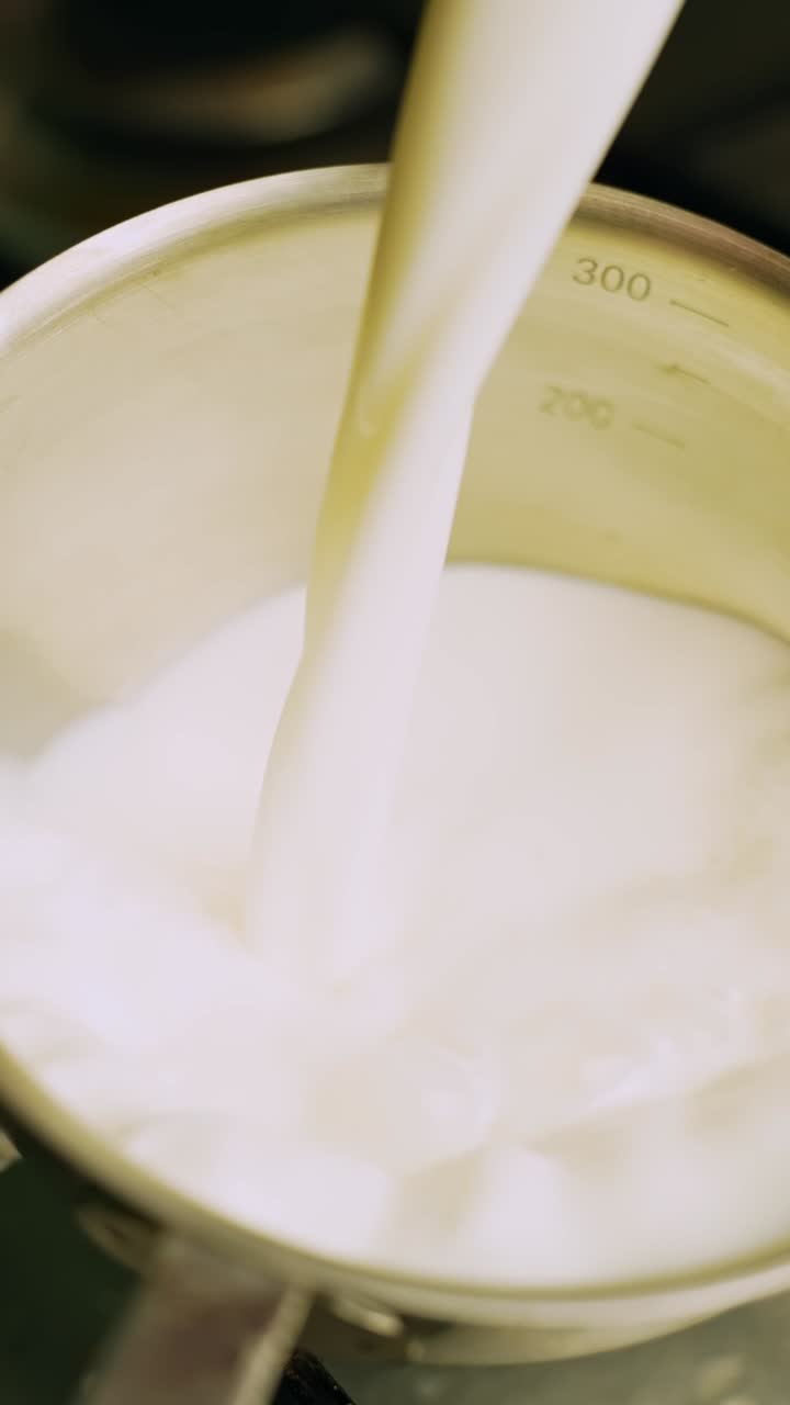 Leche vertiéndose en una cacerola de acero inoxidable en la estufa, primer plano de la preparación de lácteos para salsa o gachas. Material de archivo vertical de cocina con espacio para texto.