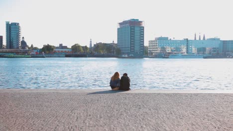 a boy and a girl sitting close to each other at the edge of a dock boulevard with the river and cityscape behind them