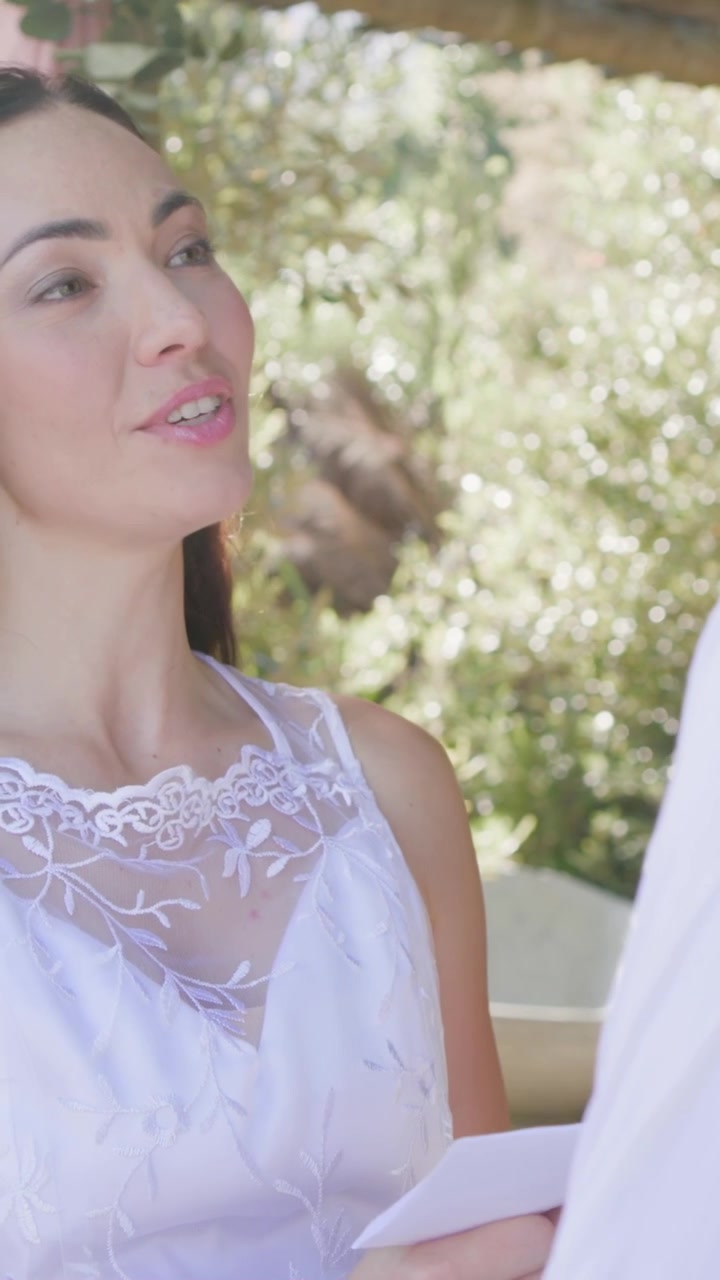 Video verticale: Donna in abito bianco che entra, legge le promesse nuziali piegate sotto un pergolato e consegna un biglietto a un uomo.