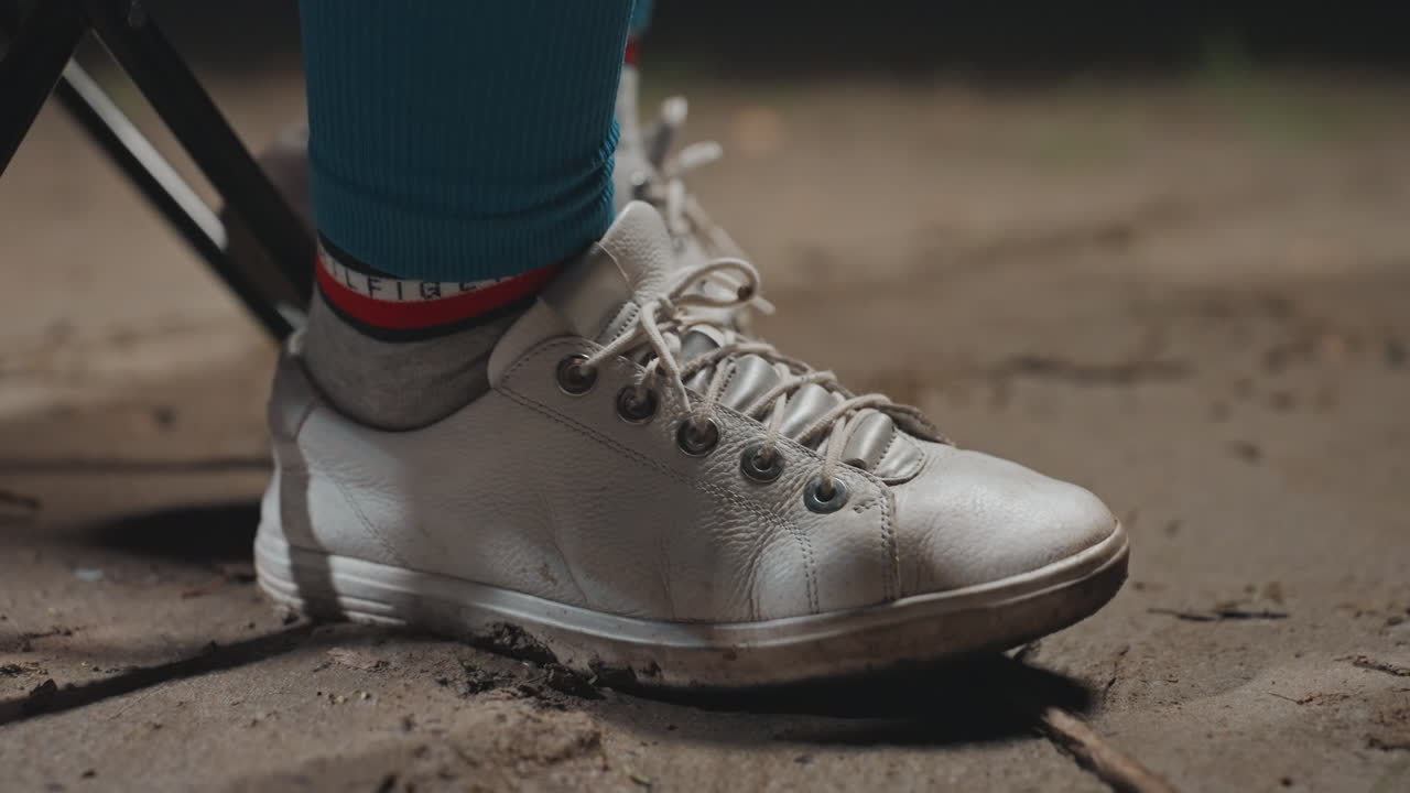 Primo piano di una scarpa da ginnastica argentata sul terreno del campeggio; lacci fangosi e calzino verde acqua visibili; la gamba di una sedia pieghevole sostiene un campeggiatore rilassato; movimento sottile mentre il piede preme nel terreno; la luce serale evidenzia la texture.