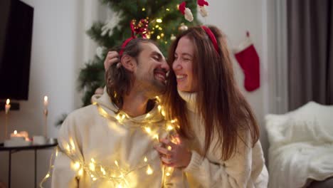 Close-up-shot-of-a-happy-brunette-girl-in-a-White-sweater-together-with-her-boyfriend-fooling-around-and-decorating-it-with-a-Christmas-glowing-yellow-garland-in-a-cozy-room-decorated-in-the-style-of-Christmas