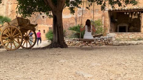 Back-view-of-ittle-girl-on-swing-with-Sicilian-cart-or-carretto-siciliano-at-Grotta-Mangiapane-or-Scurati-Caves-in-Sicily,-Italy