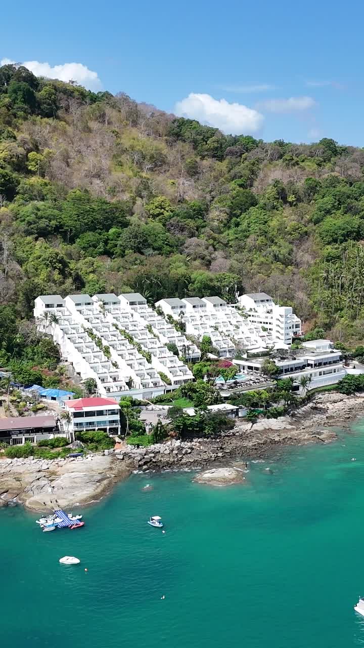 드론 영상은 태국 푸켓의 조용한 해안선을 포착합니다. 푸른 초록색, 푸른 물, 그리고 은 하늘 아래 고정된 배를 보여줍니다.