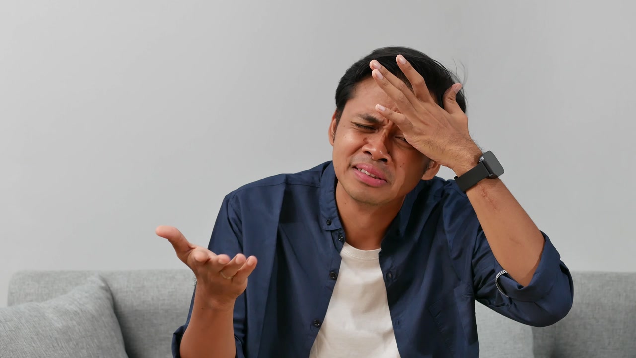 Confused young asian man sitting on sofa at home with asking hand gesture for choice, option and question