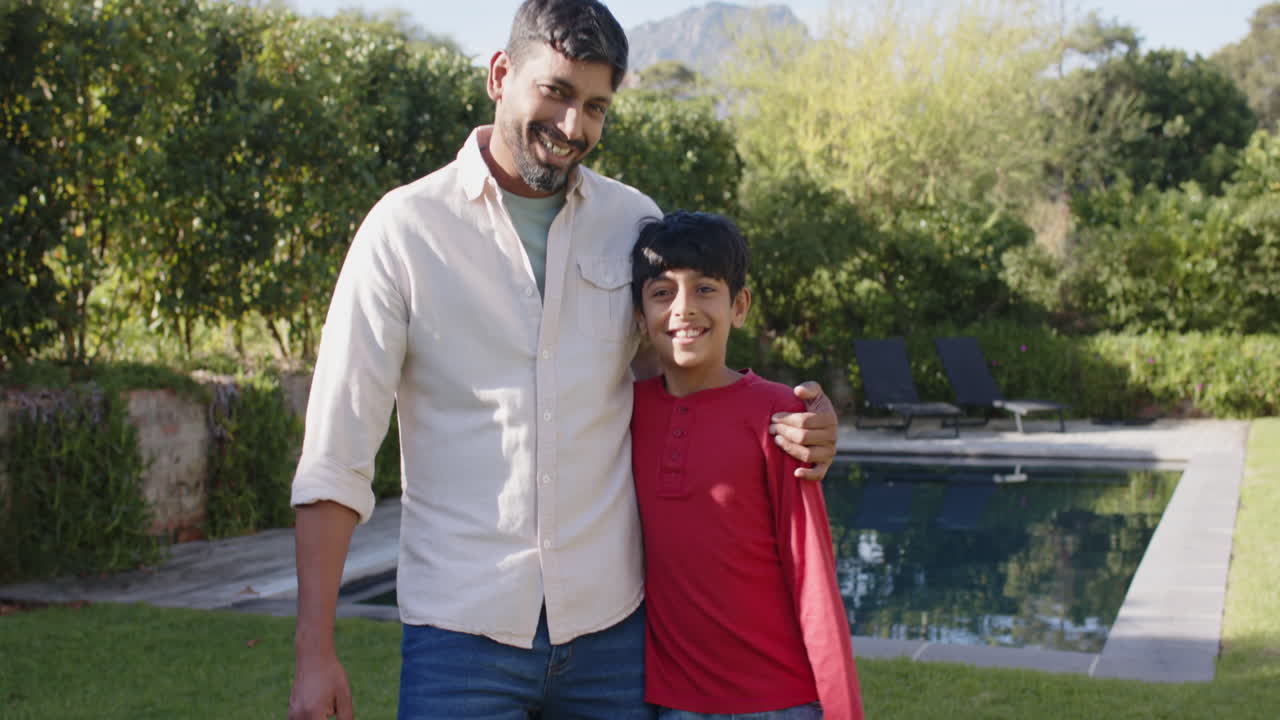 Smiling Indian father and son standing together by swimming pool in backyard