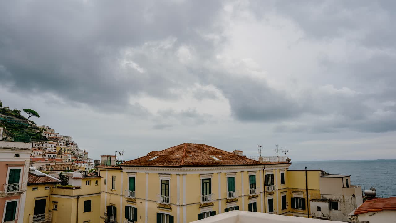 Timelapse di nuvole temporalesche sopra i tetti della Costiera Amalfitana con cielo scuro e drammatico, orizzonte marino e villaggio collinare. Atmosfera cinematografica da travel reel italiano.