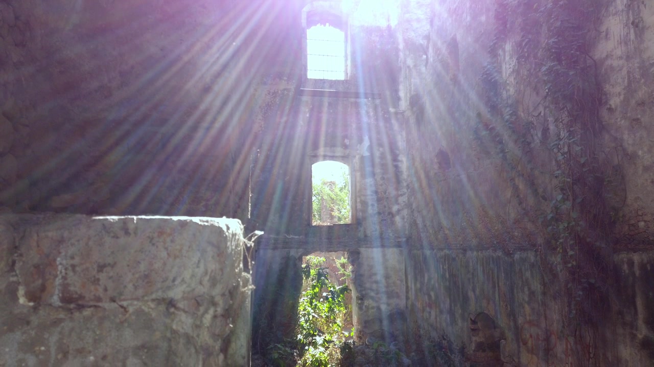 Luce solare che filtra attraverso un'apertura della finestra all'interno di un'hacienda abbandonata, raggi che illuminano le pareti in pietra e le piante che crescono all'interno della struttura, luce e ombra drammatiche in una rovina storica