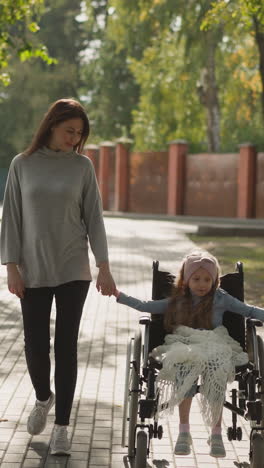 smiling mother and daughter with paralysis in wheelchair enjoy walk in park. woman spends time with little girl on fresh air on sunny spring