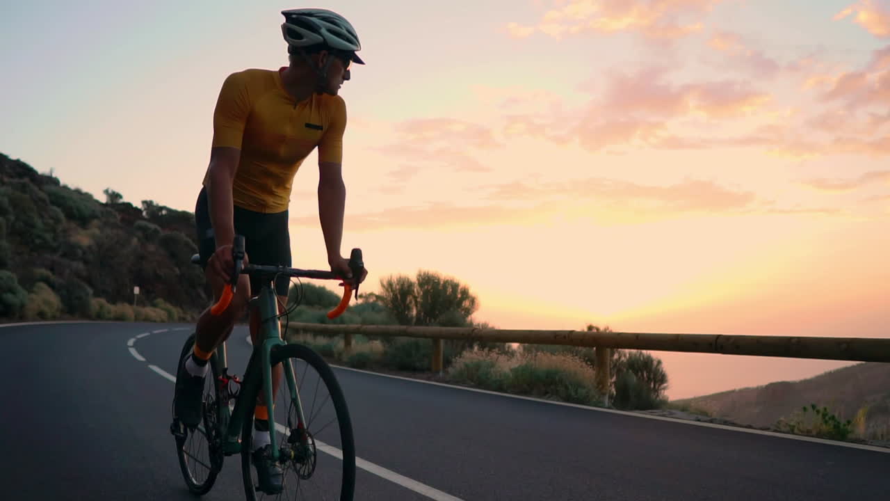 Slow motion footage depicts the athlete on a bike traversing a mountain serpentine, appreciating the island's view, embodying dedication to a healthy lifestyle at sunset