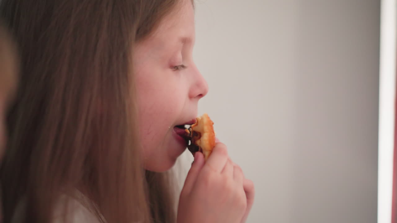 Close-up of young girl savoring chocolate-filled cookie, with partial view of another person nearby, enjoying their snack in relaxed indoor setting