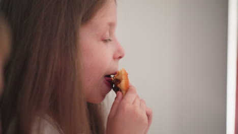 close-up of young girl savoring chocolate-filled cookie, with partial view of another person nearby, enjoying their snack in relaxed indoor setting