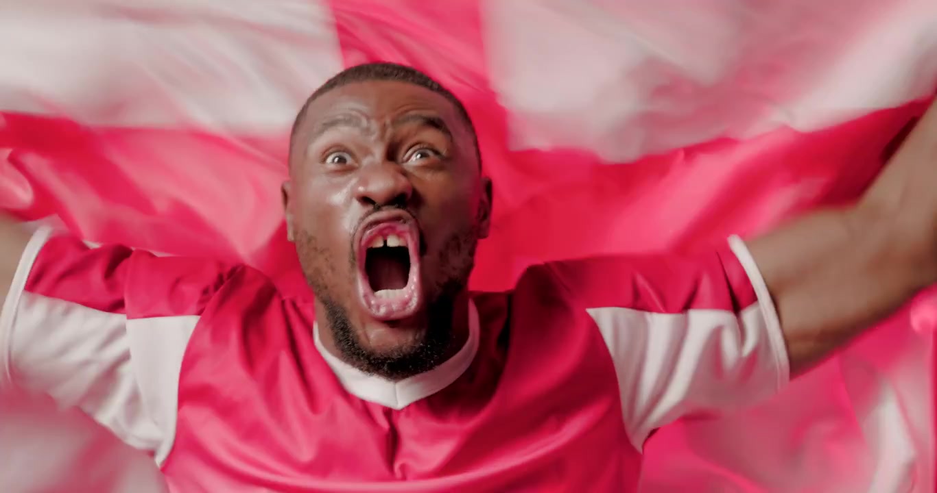 African man wearing red jersey, lifting red white flag, waving, shouting, cheering on red backdrop