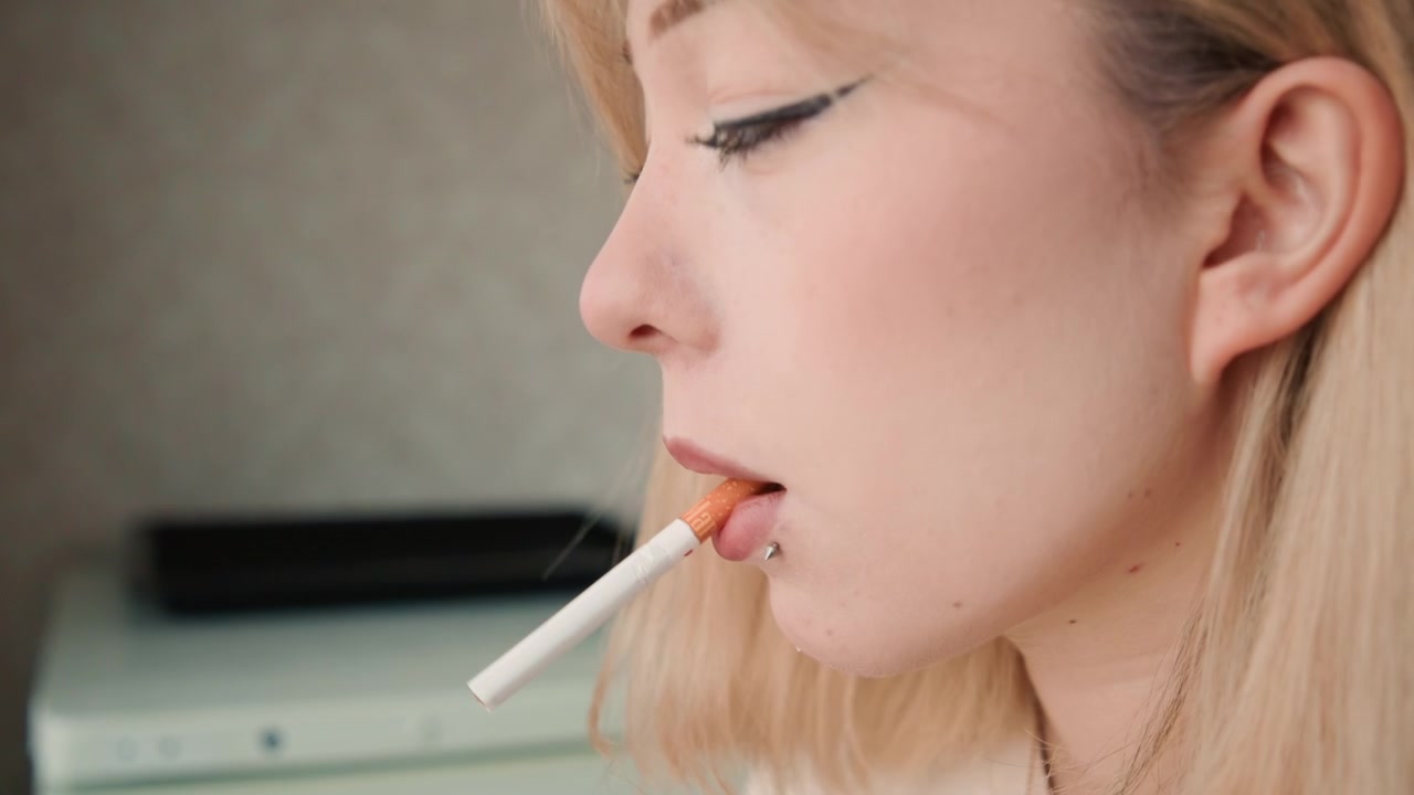 Mujer pensativa con cigarrillo en la cocina. Mujer contemplativa de cabello rubio fumando cerca. Mujer caucásica pensativa de cabello rubio y piercing labial sosteniendo un cigarrillo cerca de un electrodoméstico de cocina.